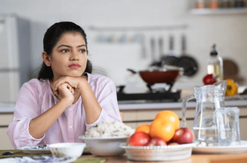 A woman sitting in a kitchen, looking thoughtful with fresh fruits and a glass pitcher on the table.