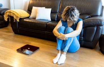 A person sitting on the floor looking discouraged after checking their weight on a scale.